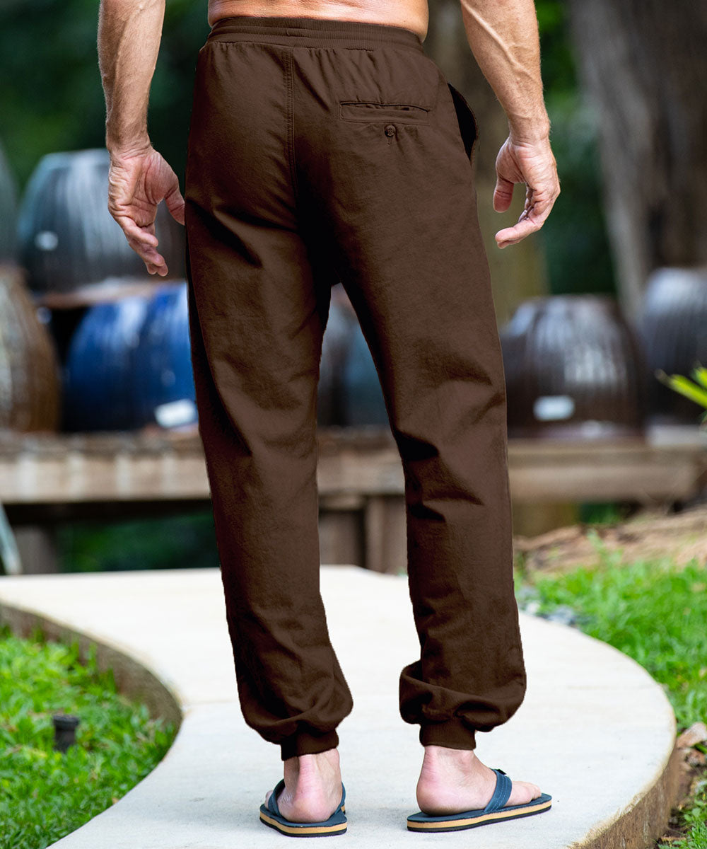 A man viewed from behind wearing light khaki linen jogger pants with elastic cuffs and a back pocket, standing outdoors on a curved concrete path with green grass and wooden barrels in the background. View 3