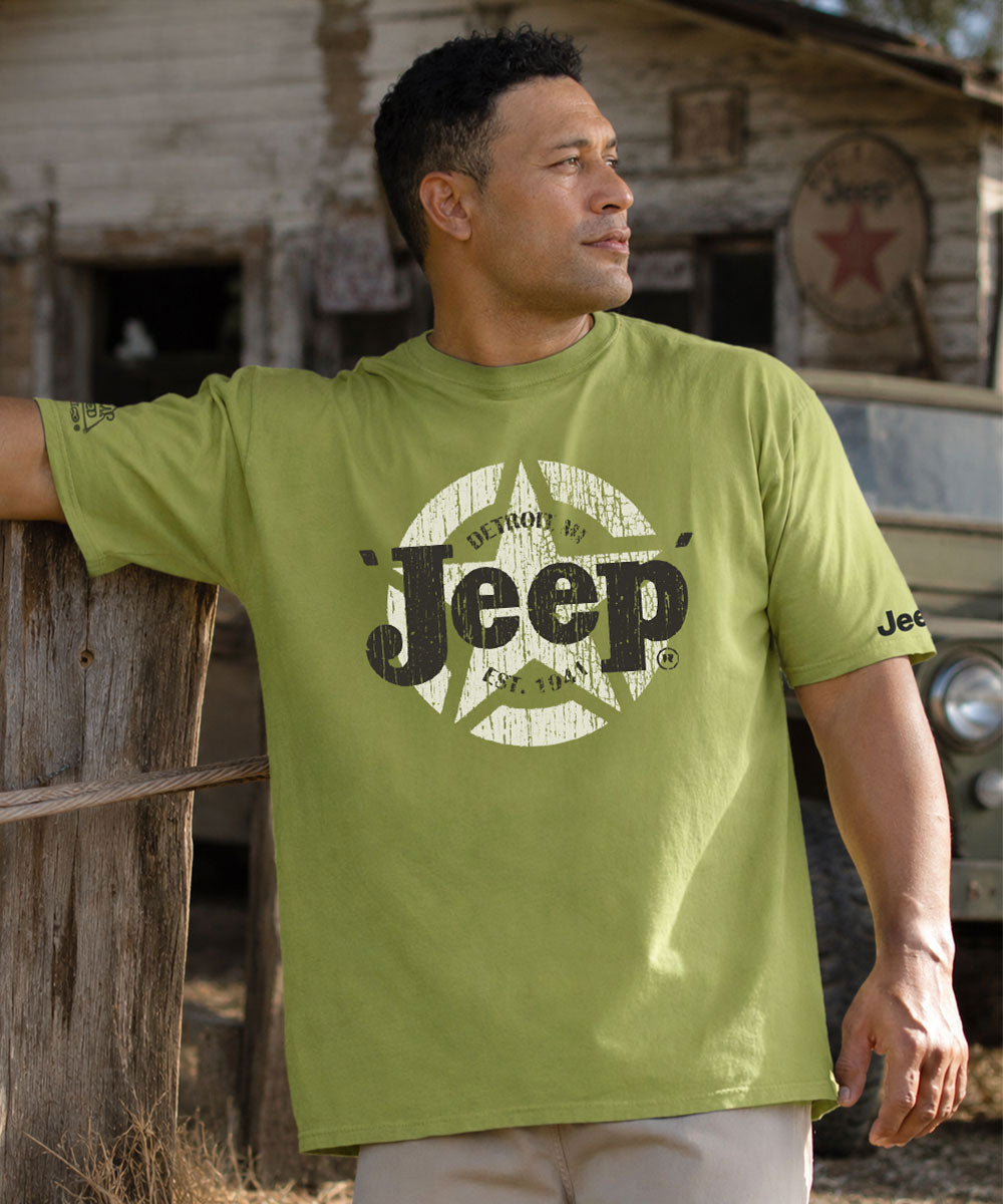 Man wearing a green Jeep t-shirt leaning against a wooden fence with trees in the background. View 2