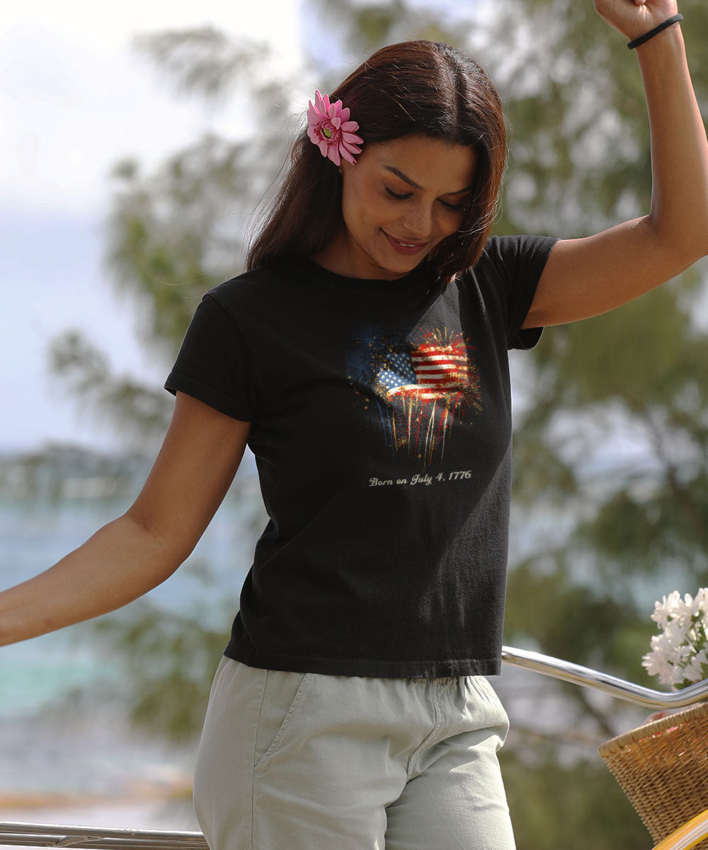 Woman wearing a black t-shirt with a graphic design, standing outdoors with trees and flowers in the background. View 2