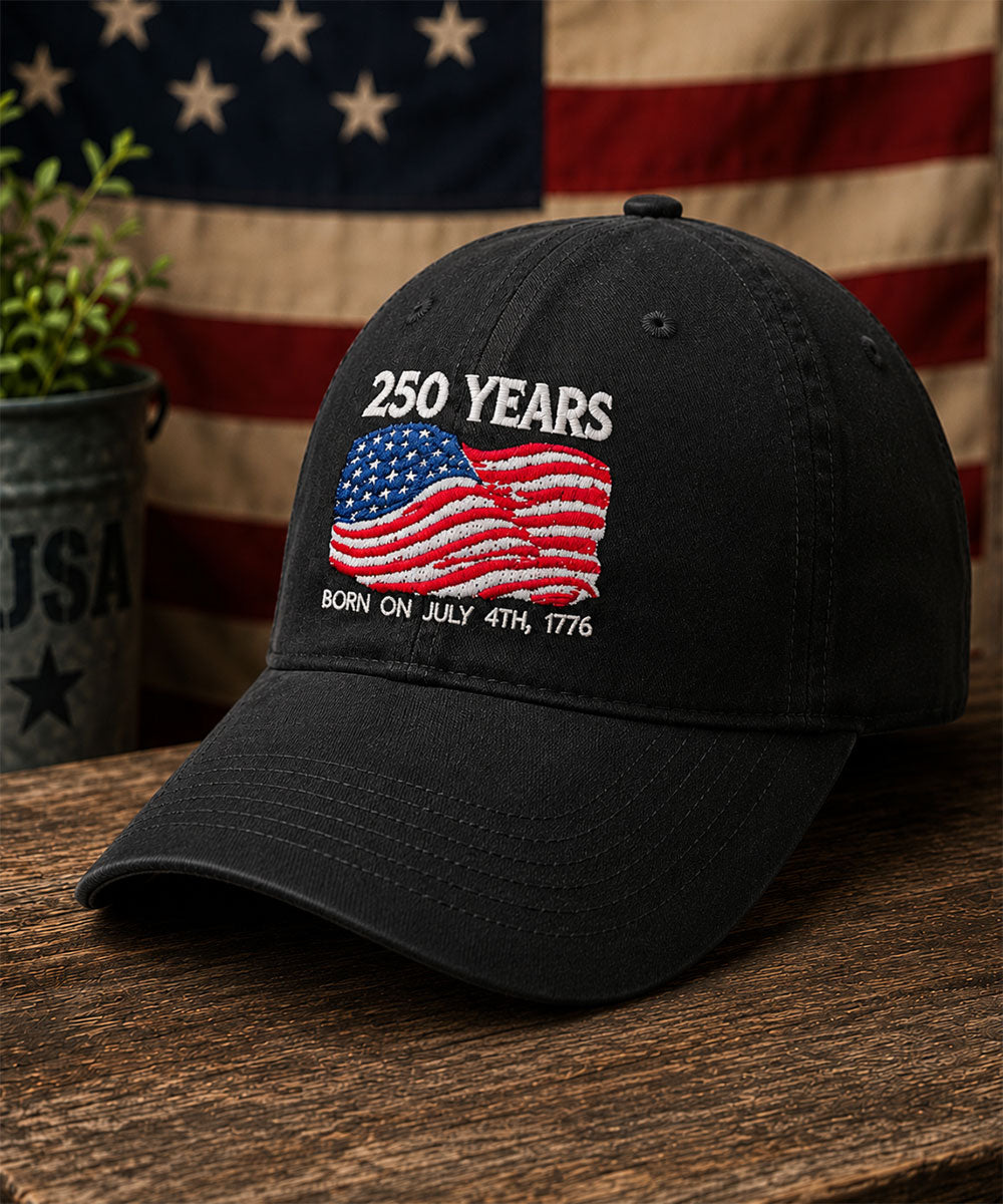Black cap with '250 Years' and American flag design on a wooden surface with an American flag in the background. View 2