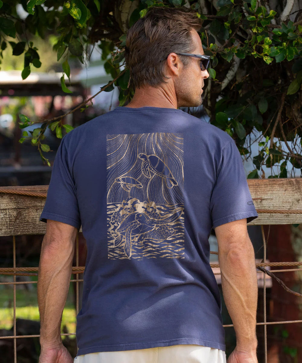 A man wearing a navy blue t-shirt with a large beige graphic print on the back, in front of a wooden fence and green foliage. View 2