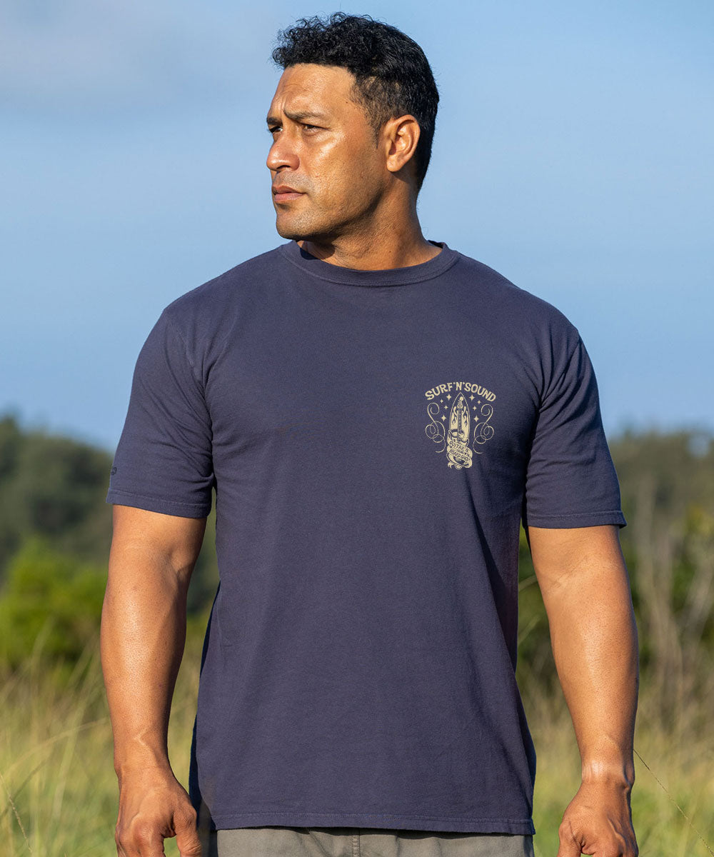 A man wearing a navy blue t-shirt with a small beige graphic on the left chest, standing outdoors in a grassy field under a blue sky. View 3