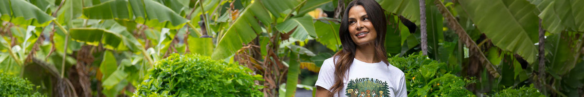 A woman with long dark hair stands outdoors in front of lush green banana leaves and tropical plants, wearing a white graphic T-shirt with a colorful circular design.