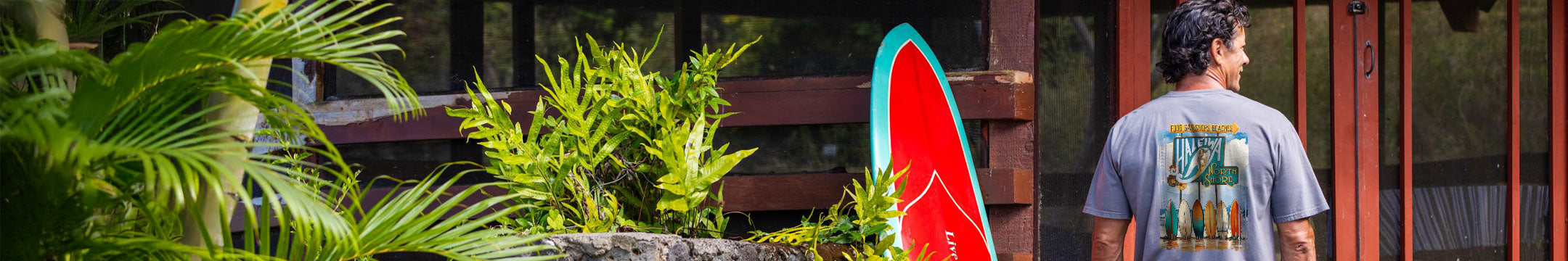 Person standing in a tropical setting with a surfboard, surrounded by greenery and a wooden structure wearing a Crater Dyed t-shirt