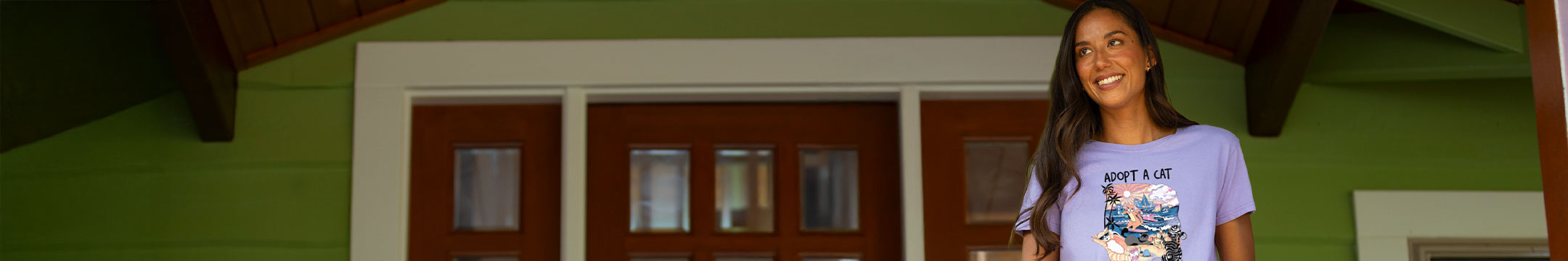A woman smiling and wearing a light purple graphic t-shirt with the text 'ADOPT A CAT' and a colorful illustration, standing in front of a green wall with wooden doors.