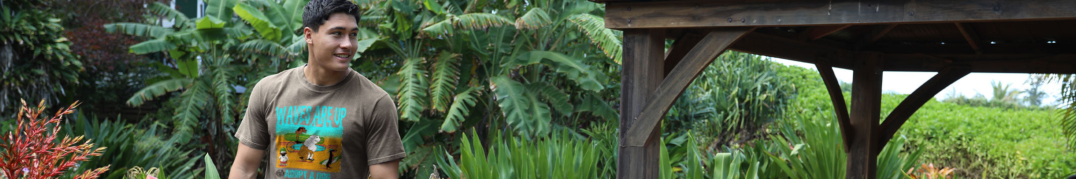 A man wearing a brown graphic t-shirt stands outdoors in a lush tropical garden next to a wooden structure.
