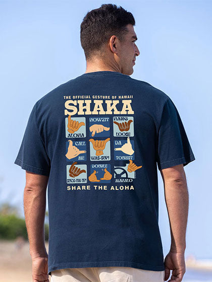A man shown from the back wearing a navy blue t-shirt with a large graphic print on the back featuring the Hawaiian word 'SHAKA' and hand gestures. The background is a blurred outdoor beach scene with blue sky.