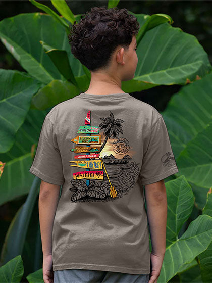 A boy viewed from behind wearing a taupe brown t-shirt with a beach graphic on the back with colorful signs, set against a background of large green tropical leaves.