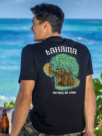Person wearing a black t-shirt with 'Lahaina' text above and our roots are strong below an outline of a tree over Maui island, standing by the ocean.