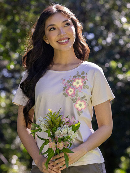 A woman outdoors wearing a cream-colored short-sleeve t-shirt with a pink and green floral graphic on the left chest, holding a small bouquet of flowers.