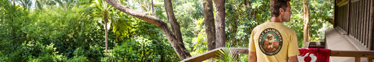 A man wearing a yellow t-shirt stands on a wooden deck overlooking a lush green tropical forest. The back of the t-shirt features a circular graphic with the print "Mele Kalikimaka"