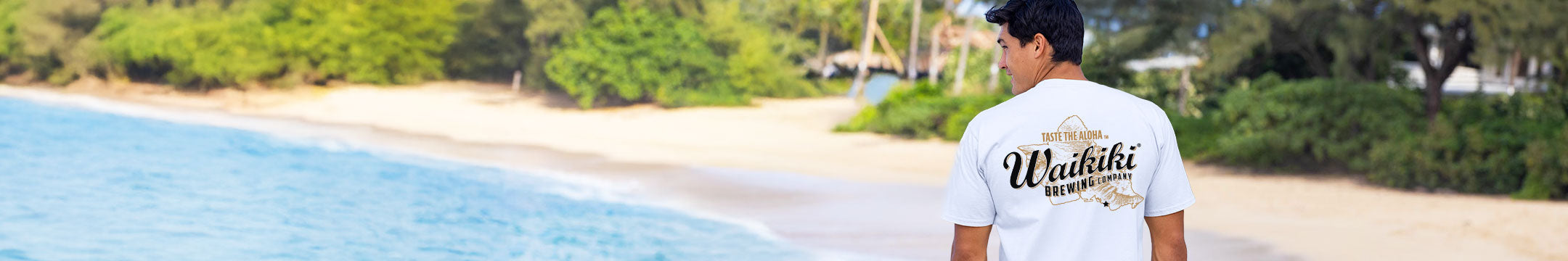 Person wearing a 'Wailiki' shirt standing on a beach with blue water and greenery.