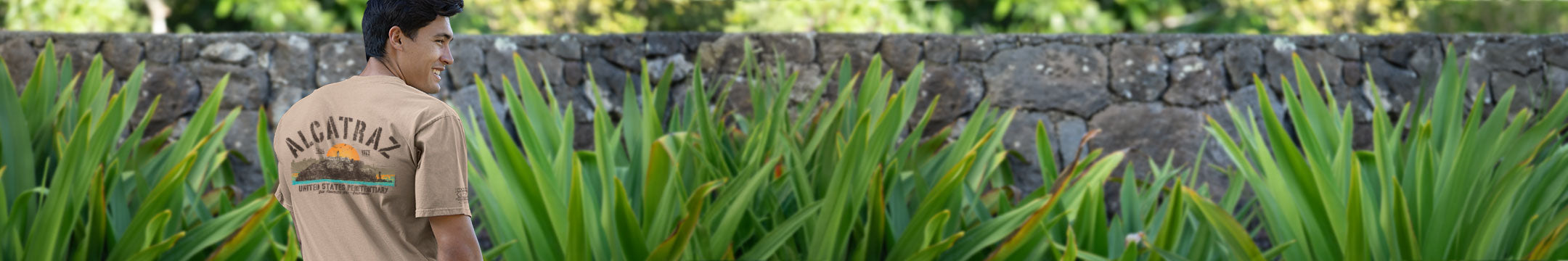Person wearing a brown shirt with 'alcatraz' logo walking through green plants.