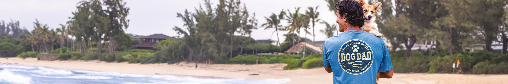 Man in a blue 'Dog Dad' shirt standing on a beach with a dog.