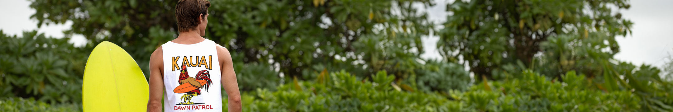 Person holding a yellow surfboard with 'Kauai' and 'Hawaii' text, surrounded by green foliage.