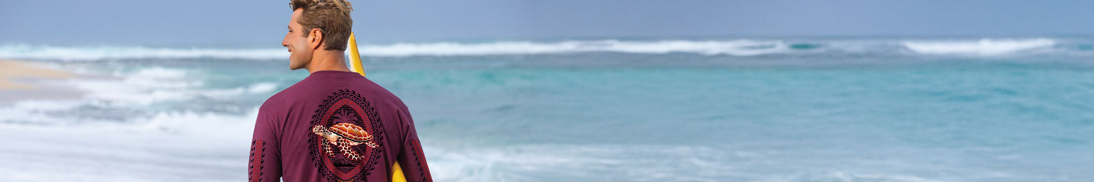Man standing on a beach looking at the ocean with a surfboard under his arm.
