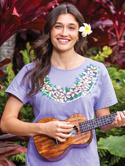 Woman playing a ukulele with a floral design on her shirt, surrounded by tropical plants.