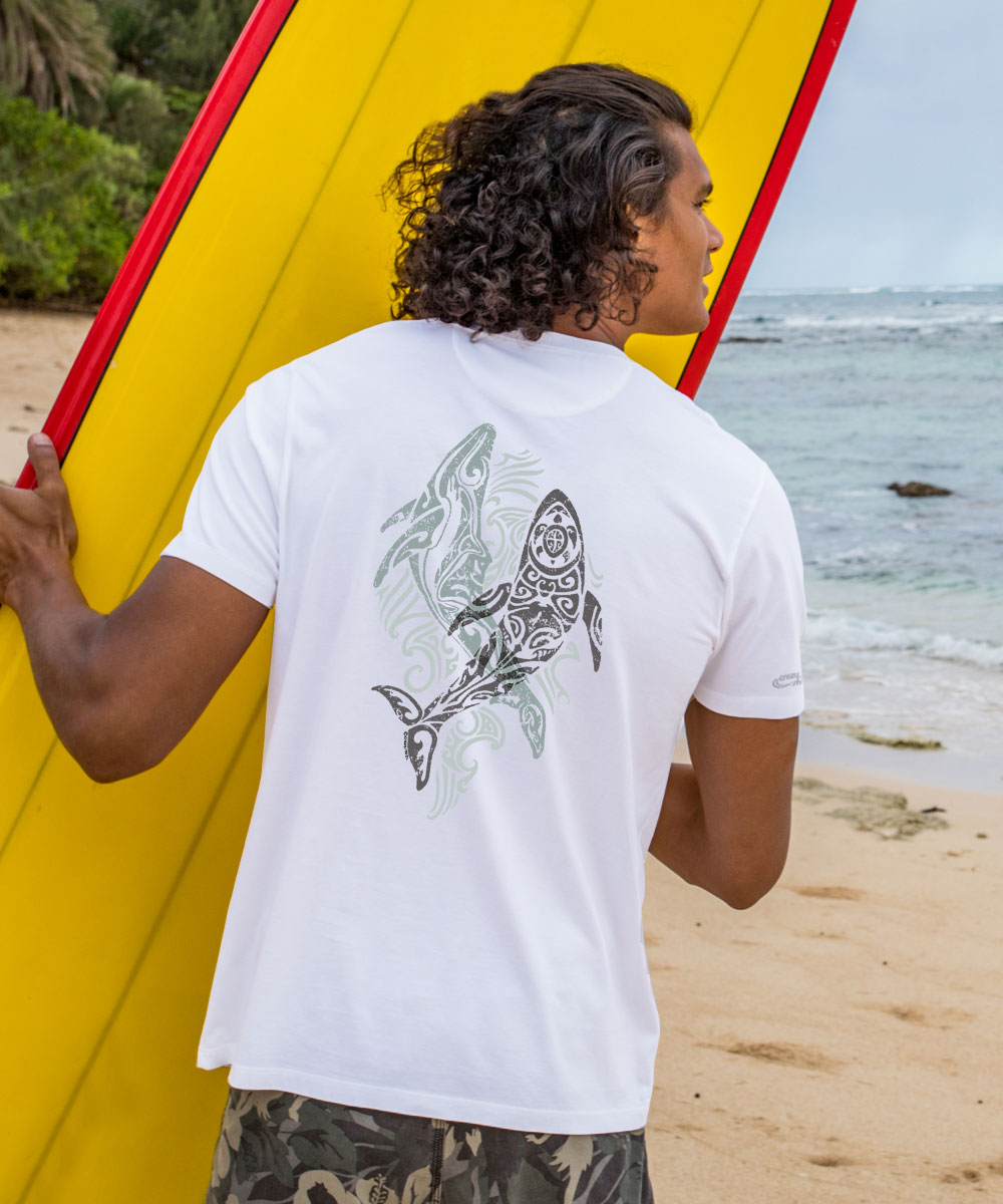 A person standing on a beach wearing a white short sleeve pima t-shirt with a tribal-style whale graphic on the back. View 2