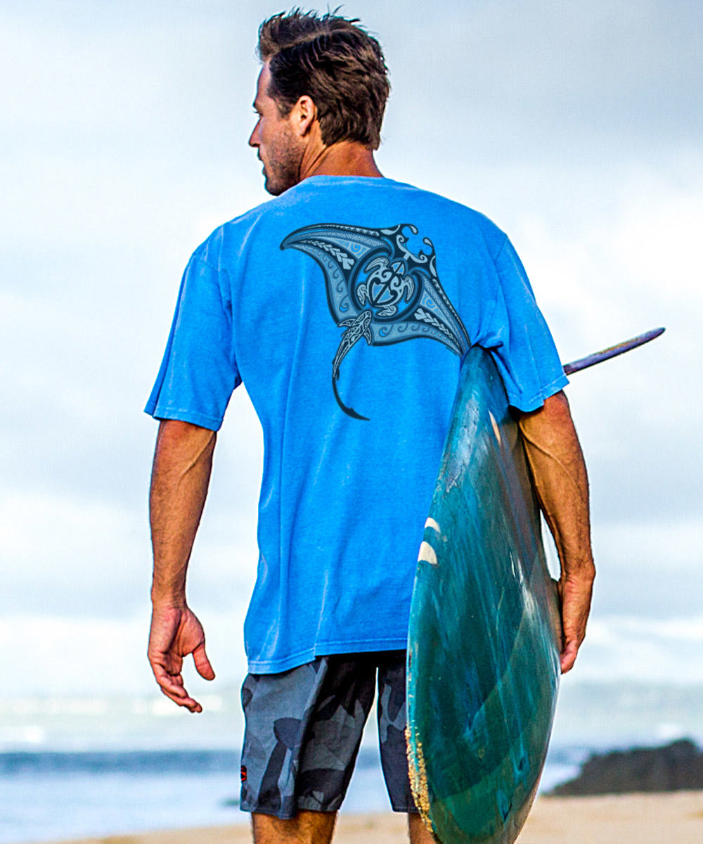 A person standing on a beach facing away from the camera, wearing a blue short-sleeve crewneck t-shirt with a manta ray design. View 2