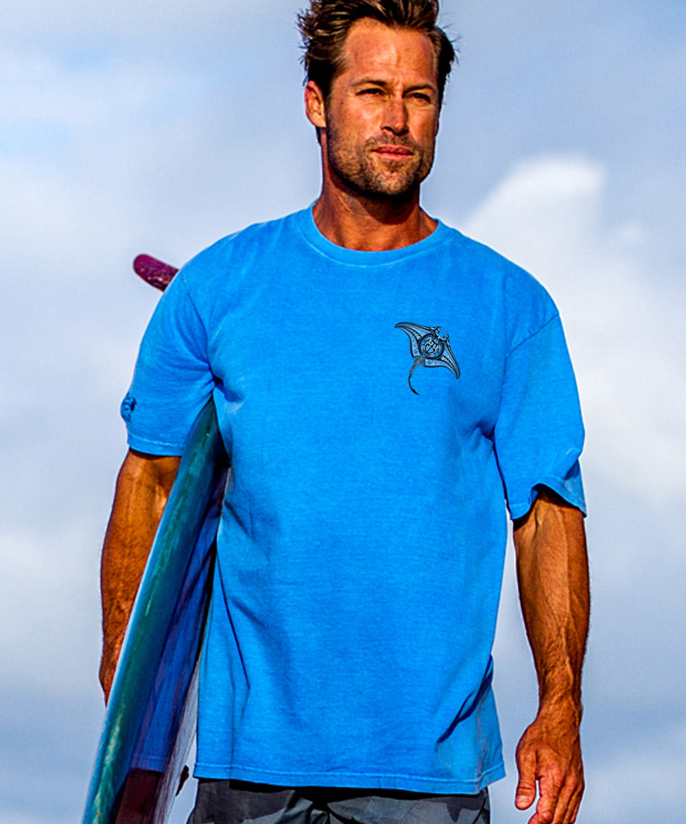 man standing on the beach holding a surfboard wearing a blue tee shirt with a logo. View 3
