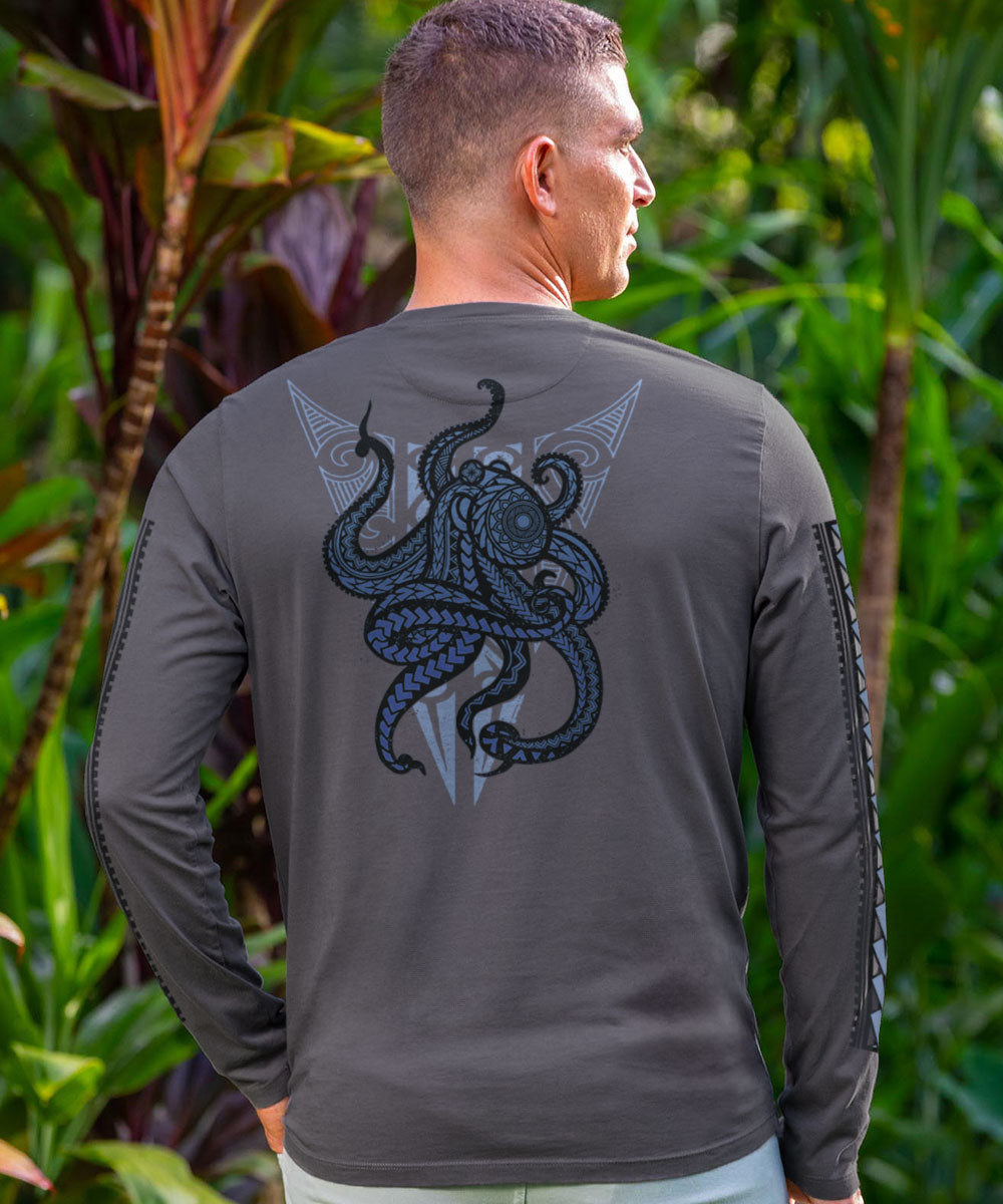 A man wearing a long sleeve gray t-shirt with a black and blue tribal octopus design on the back, standing in front of lush greenery. View 2
