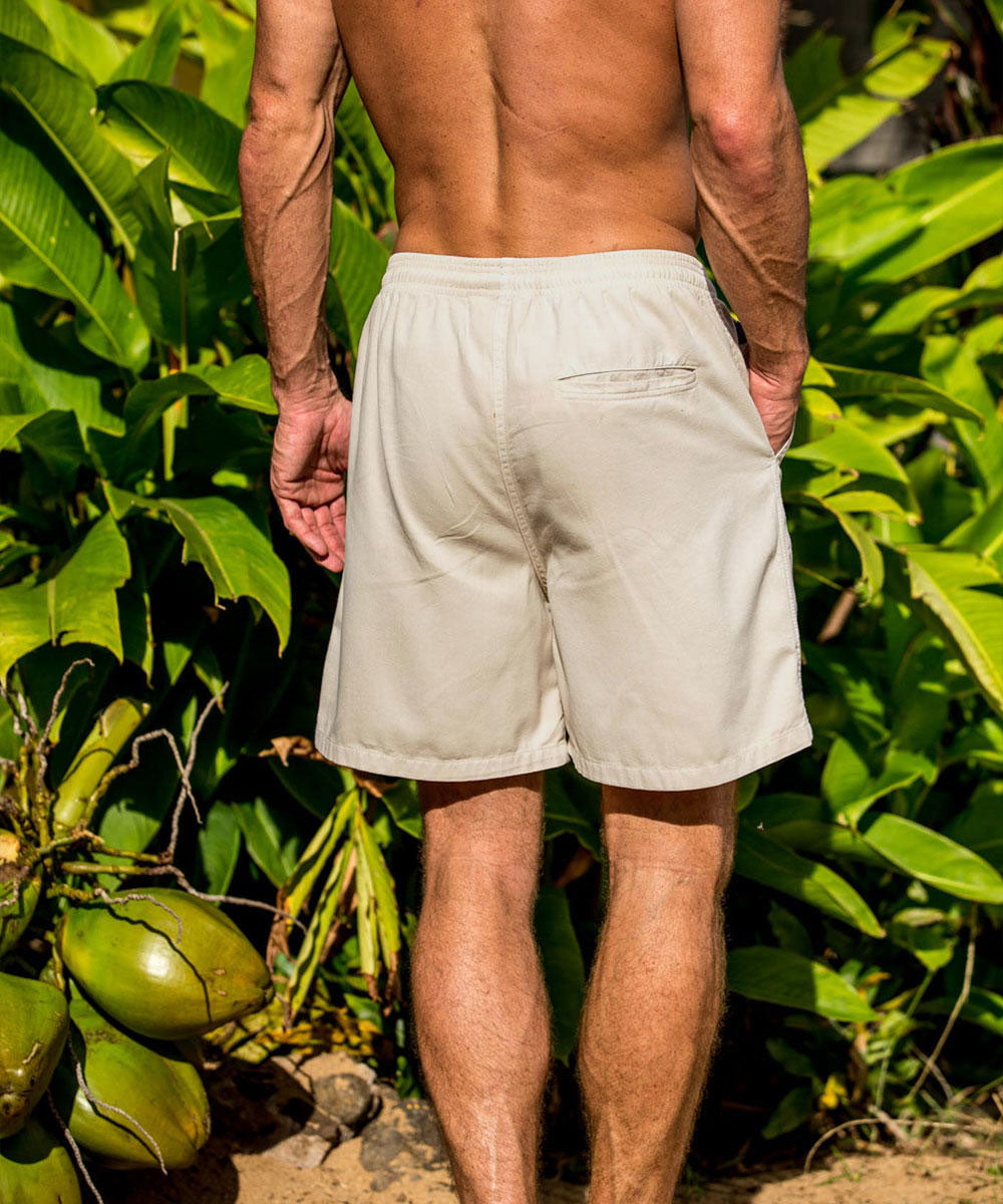 A pair of beige, twill shorts modeled by a person standing in front of a yellow wall and greenery. View 3