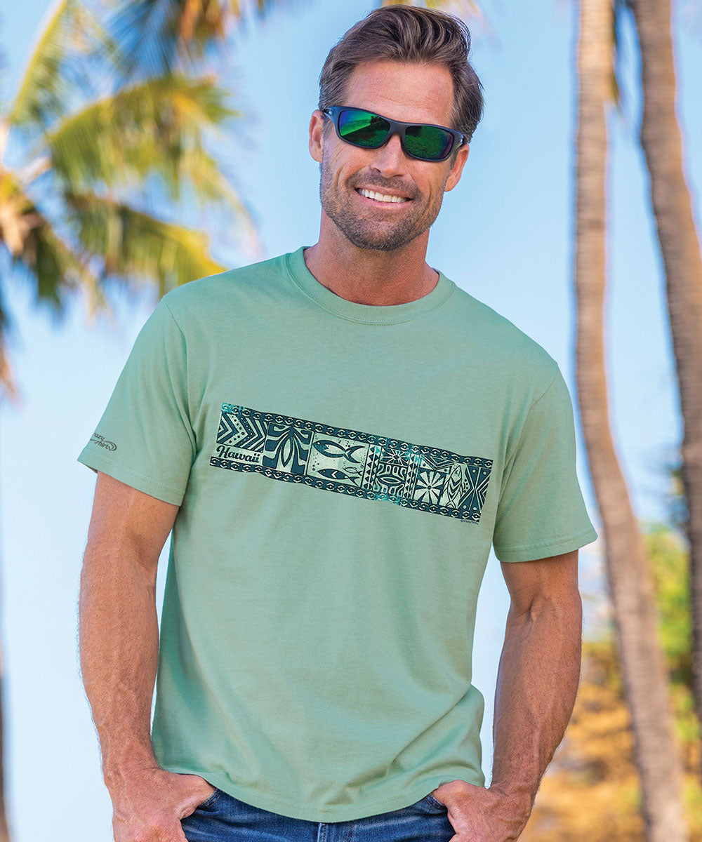 A man wearing a green short sleeve crewneck t-shirt with a geometric band design on the front, standing against a tropical backdrop with palm trees. View 2