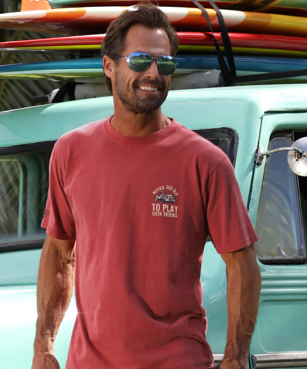 Man wearing a red short sleeve shirt with a smaller logo of the wording "Never Too Old To Play with Trucks". View 3