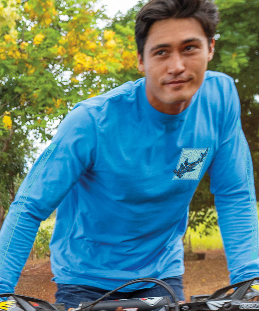 A man wearing a blue long-sleeve shirt with a small tribal hammerhead shark graphic on chest, riding a motorcycle through wilderness. View 3