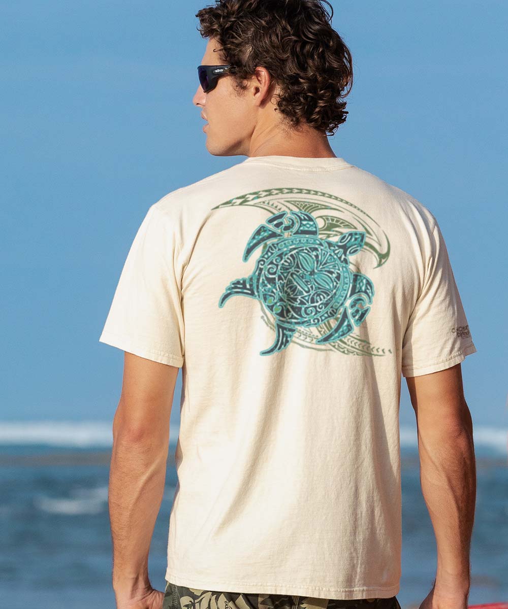 A person wearing a beige short sleeve crewneck t-shirt with a tribal sea turtle design in blue and green on the back. View 2