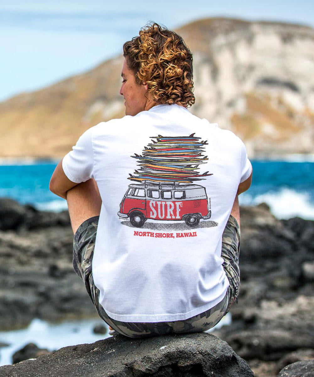 A person sitting on rocks by the beach wearing a white short sleeve crewneck T-shirt with a graphic of a surfmobile and the text 'Surf North Shore Oahu Hawaii'. View 2