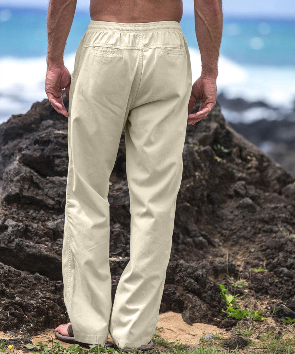 A person standing in front of a rocky outcrop wearing beige twill pants with an elastic drawstring waistband and pockets, paired with a white shirt and flip-flops. View 3
