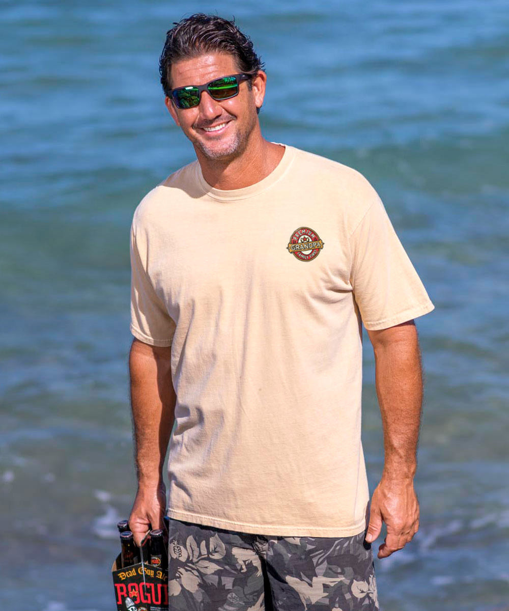 man wearing a beige short-sleeve crewneck t-shirt with the small text 'Premium GRANDPA' on it, set against a beach backdrop. View 3