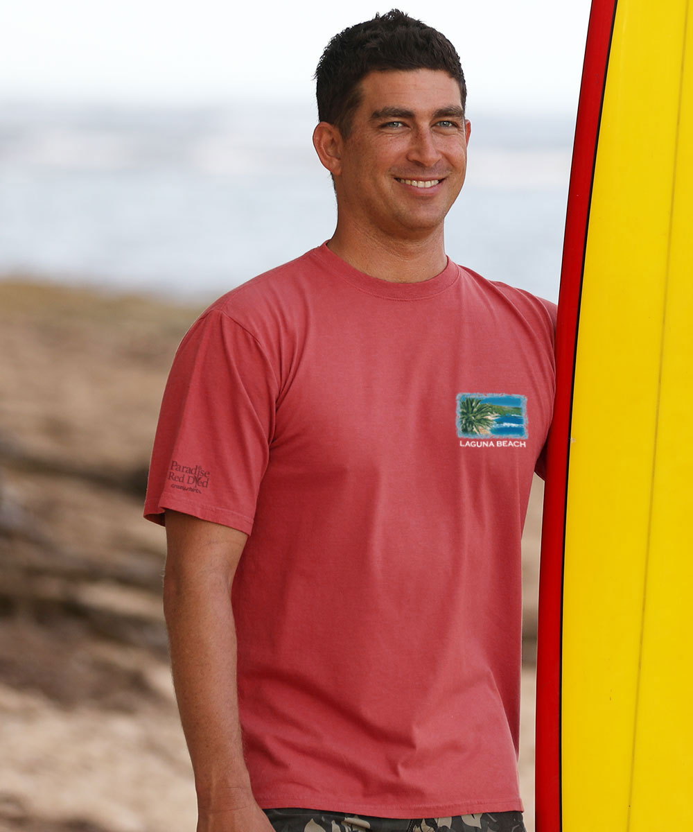Person facing towards the camera wearing a red Laguna Beach logo tee holding a surfboard View 3