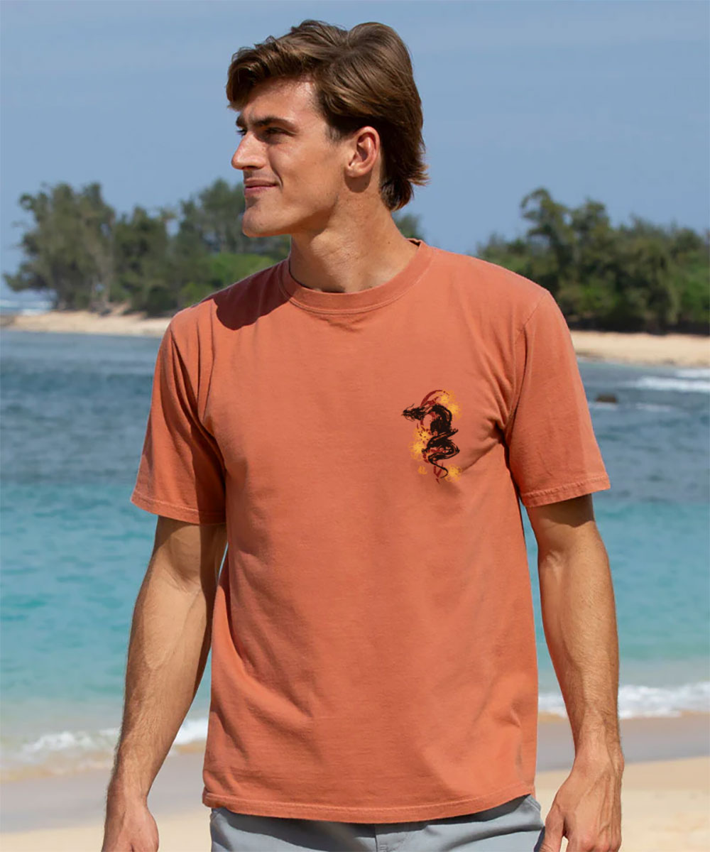 A man wearing a light red short sleeve crewneck t-shirt with a black and gray dragon design small logo on chest with the beach in the background. View 3