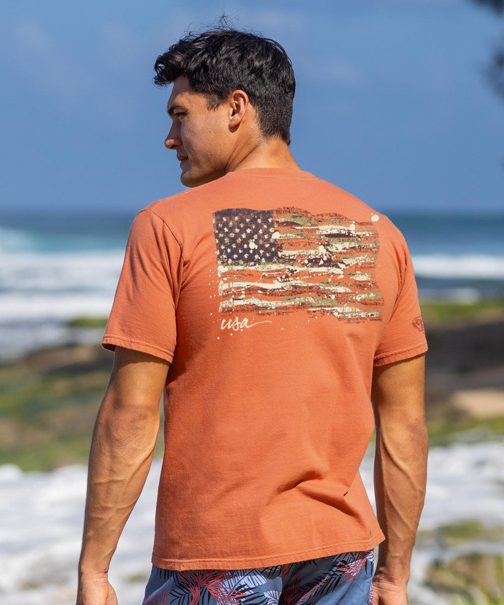 A man wearing a short sleeve crewneck t-shirt in light red with a painted American flag design on the back. View 2