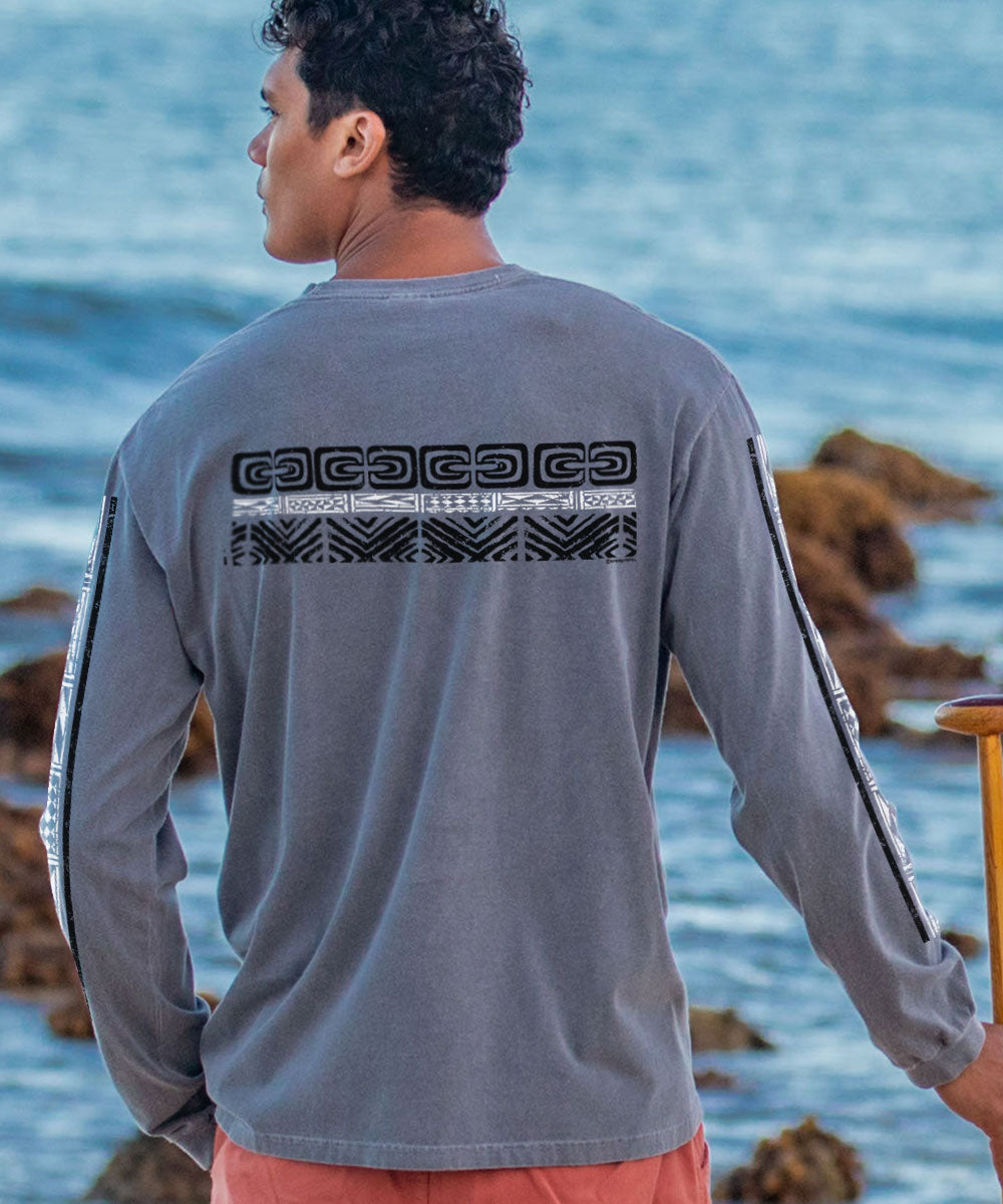 A man wearing a long sleeve crewneck t-shirt with tribal patterns printed across the back and sleeves, looking out at the sea. View 3