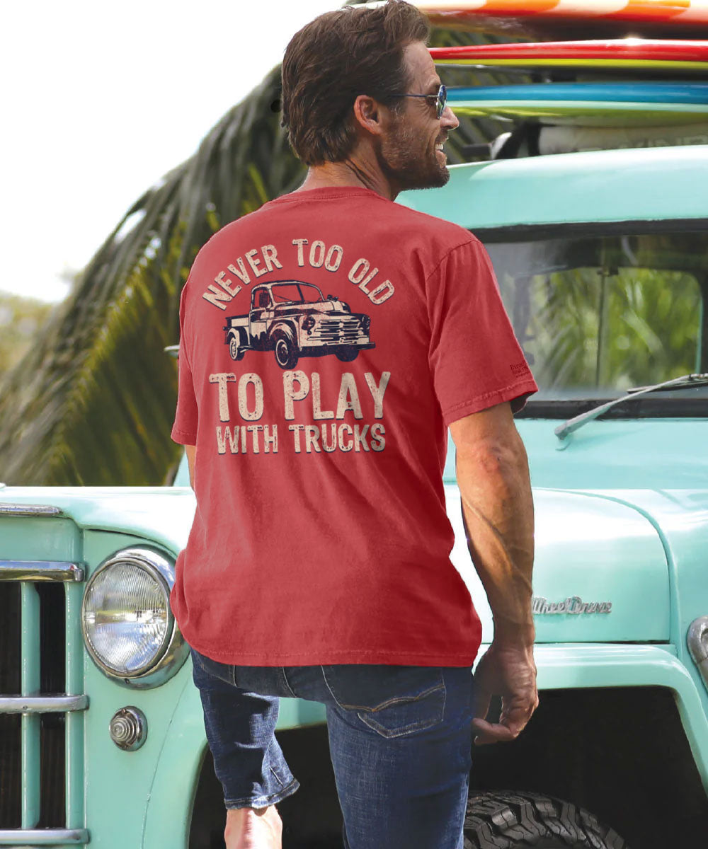 A man standing beside a vintage truck wearing a red short sleeve crewneck t-shirt with a graphic that includes a truck and the phrase 'NEVER TOO OLD TO PLAY WITH TRUCKS'. View 2