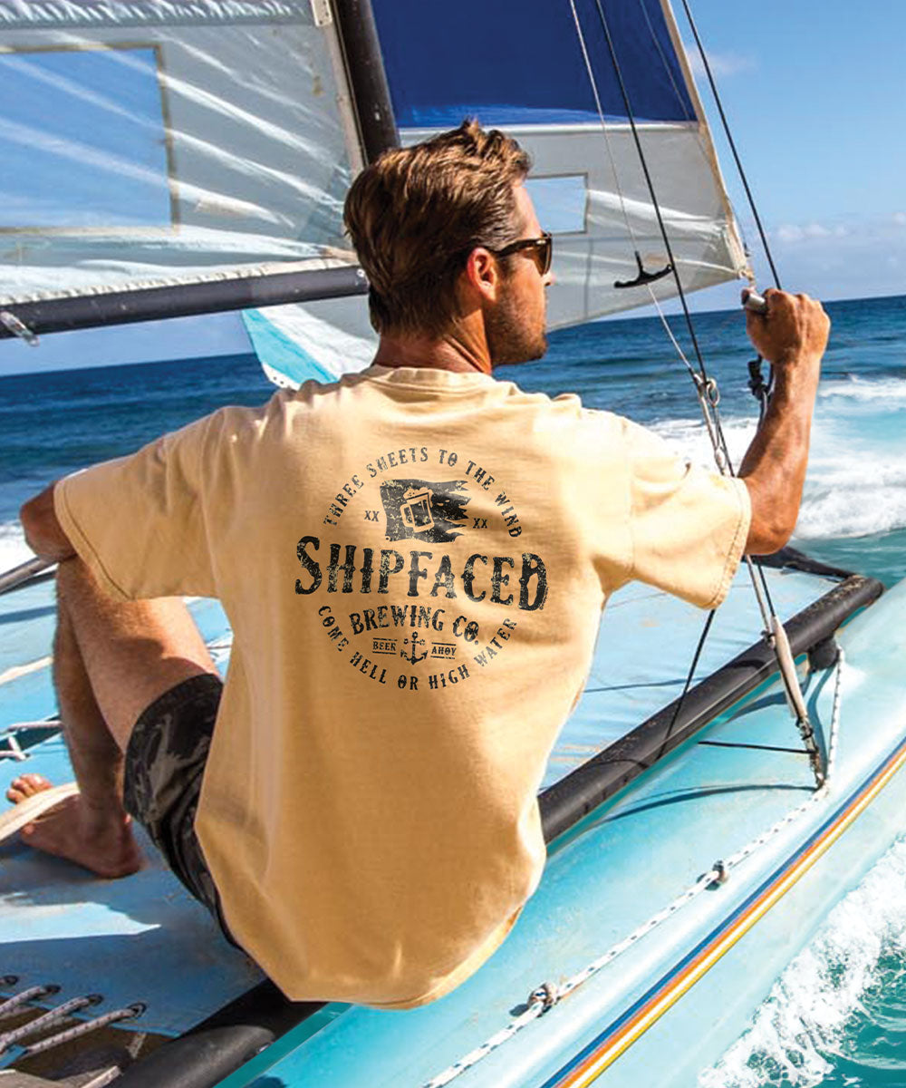 A person on a sailboat wearing a beige short sleeve crewneck t-shirt with the text 'SHIPFACED BREWING CO' and a beer glass graphic.