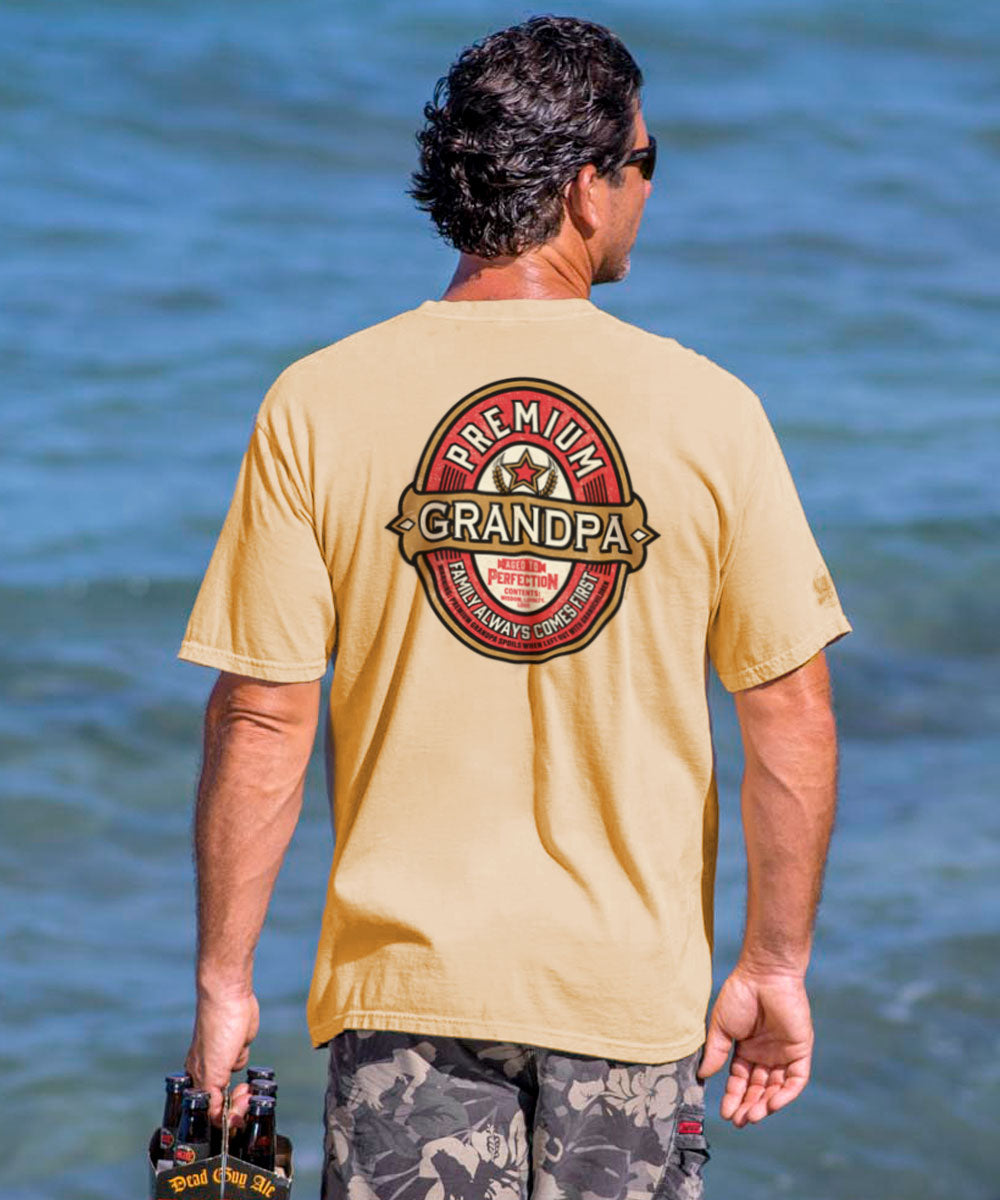 A man facing away from the camera wears a beige short-sleeve crewneck t-shirt with the text 'Premium GRANDPA' on it, set against a beach backdrop. View 2