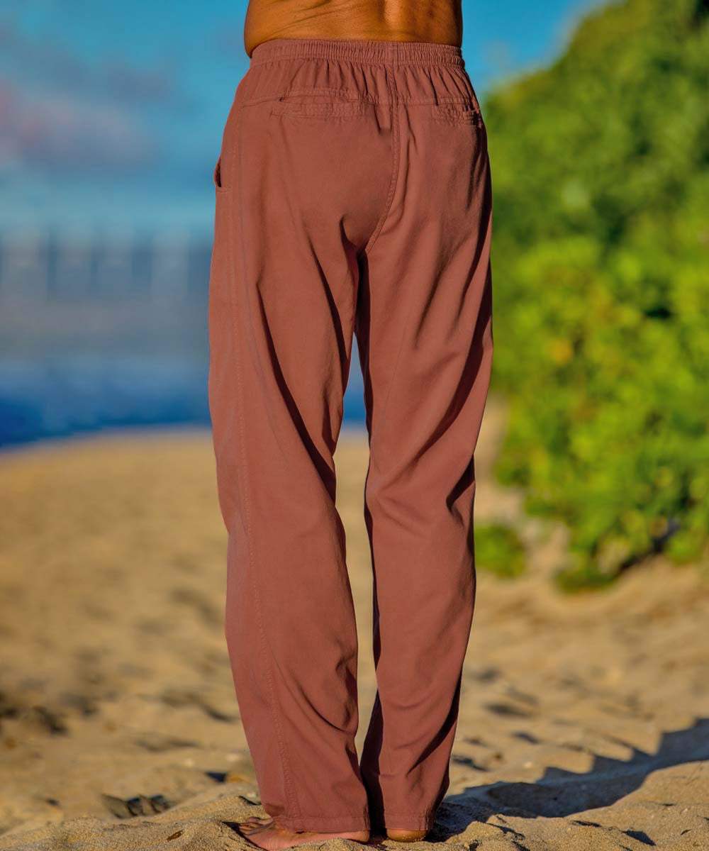 A person standing on a beach wearing rust twill pants featuring the back pockets. View 3