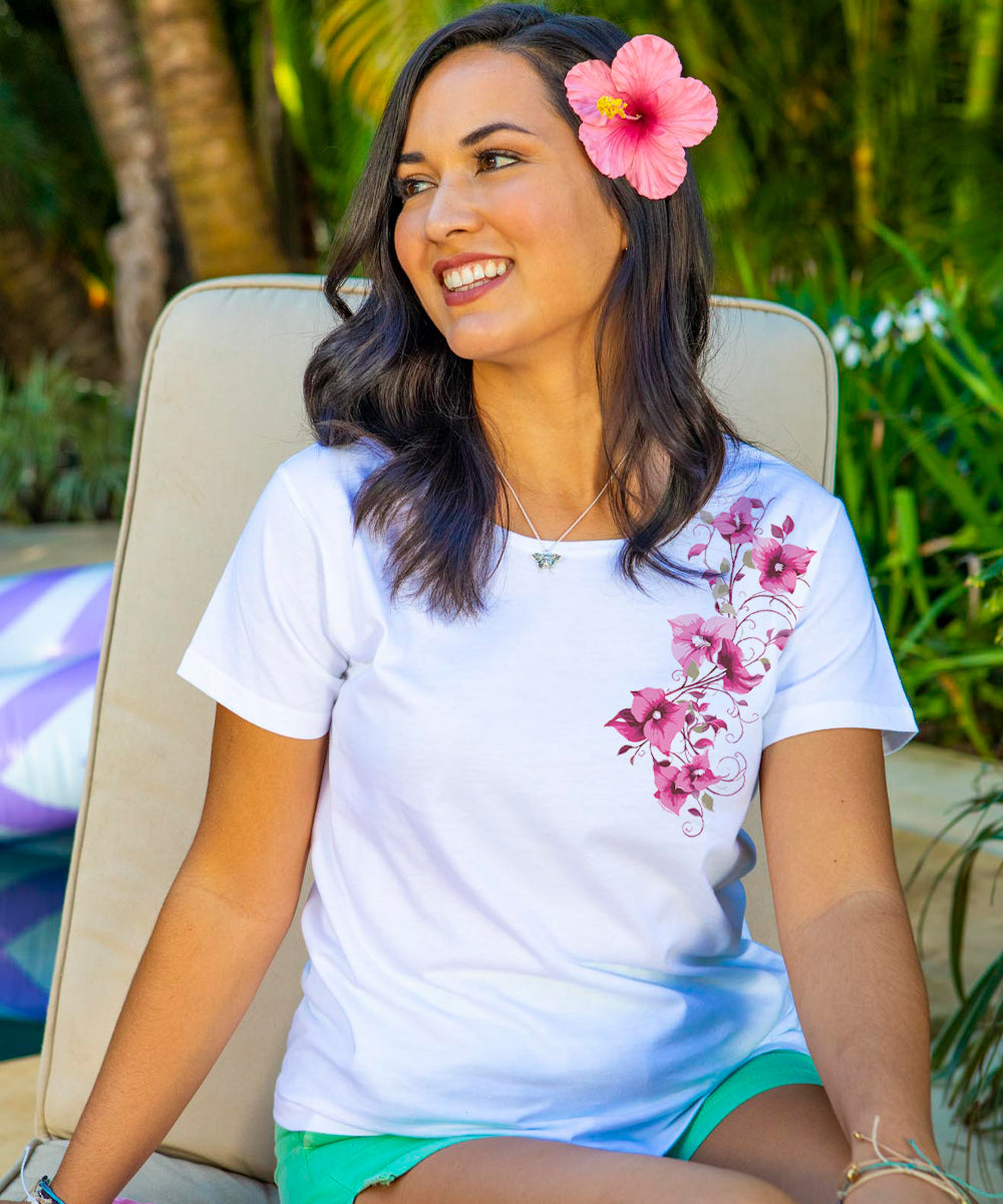 A woman sitting and smiling wearing a white short sleeve scoop neck t-shirt with a Bougainvillea floral design cascading down from left shoulder on the front. View 2