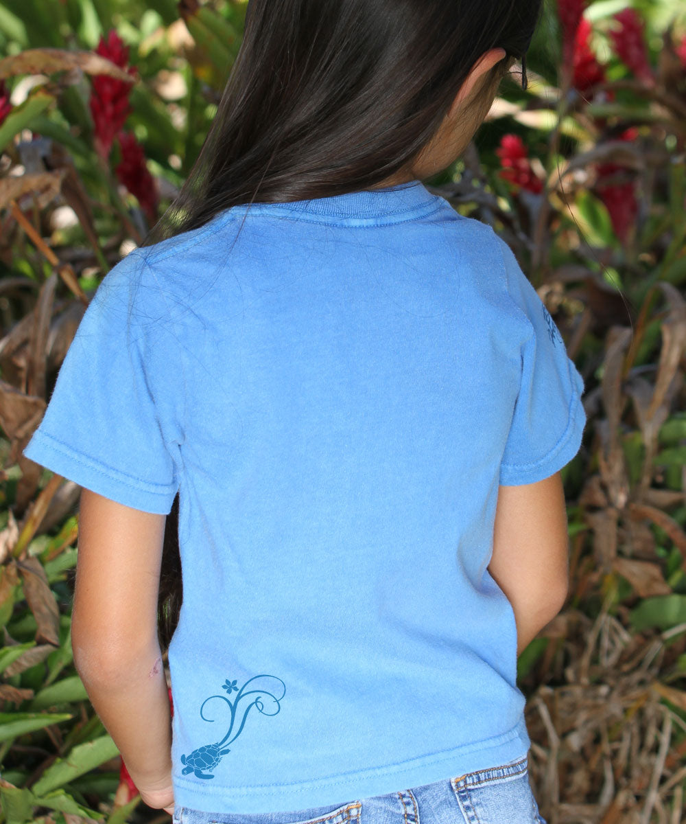 A child wearing a blue short sleeve crewneck t-shirt with a graphic design, standing in front of a tropical plant backdrop. View 3