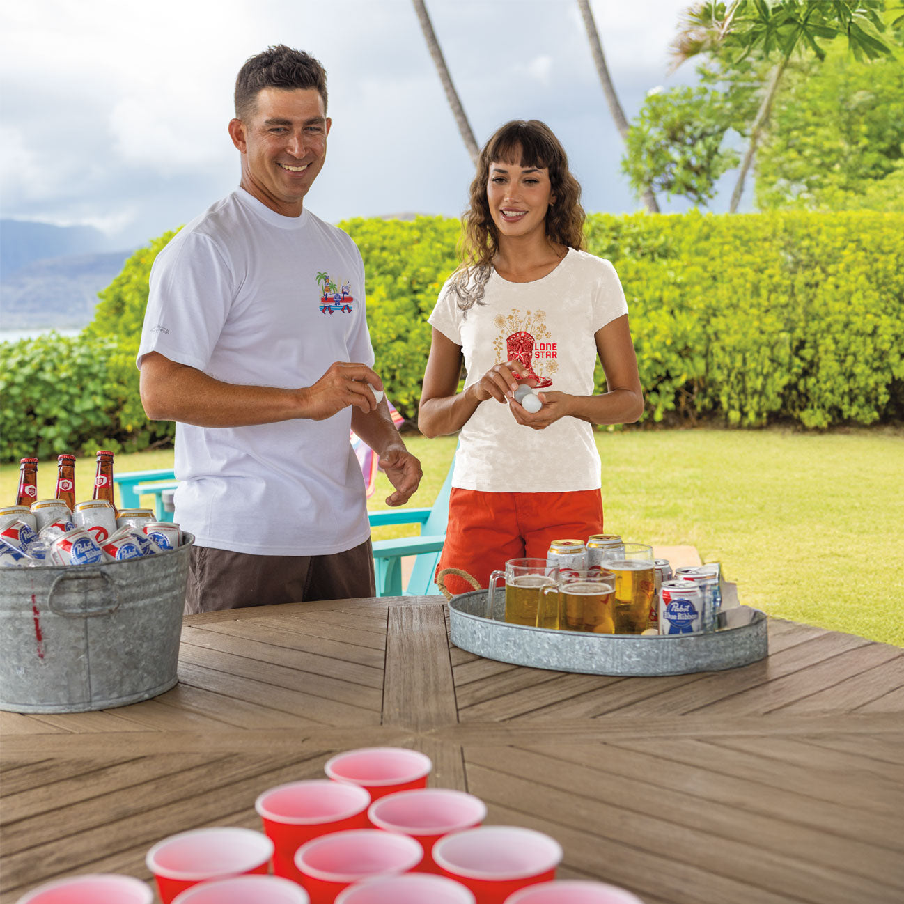 Two people standing outdoors with a table full of drinks and beer bottles.