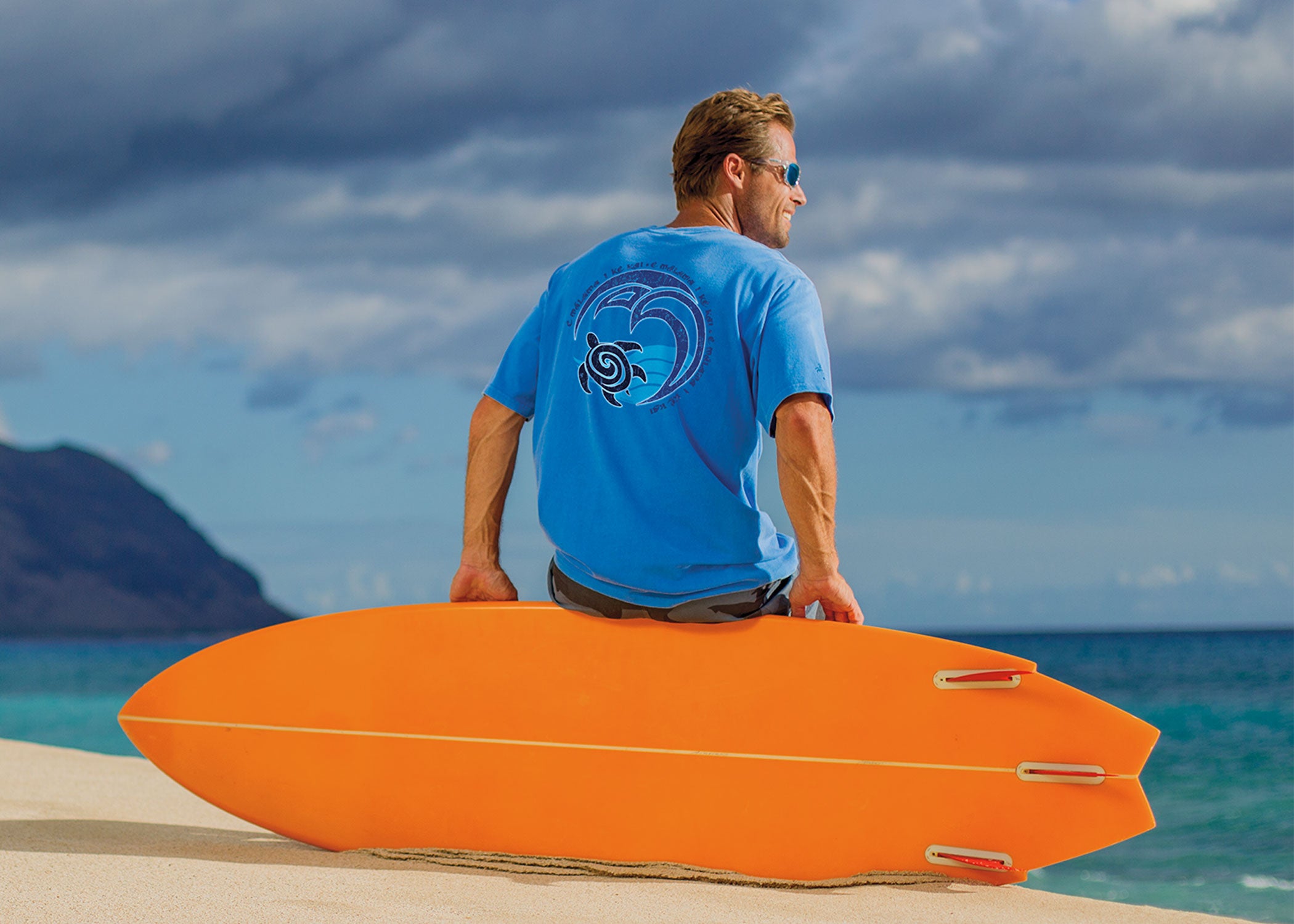 Man sitting on the edge of a surfboard on the beach looking out at the ocean