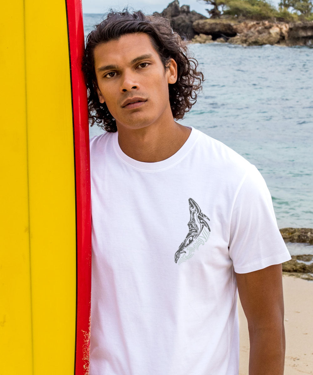 Man wearing a White t-shirt with a small black and gray graphic leaning on a colorful surg board with the beach in the background. View 3