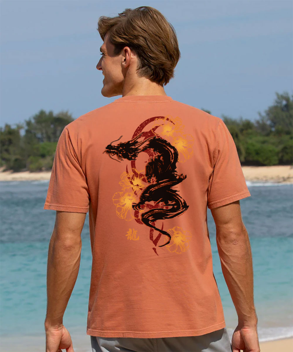 man wearing a light red short sleeve crewneck t-shirt with a black and gray dragon design on the back, standing near a beach. View 2