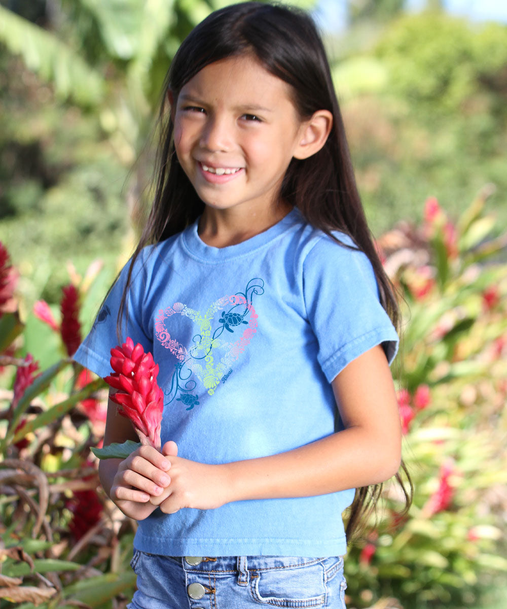 A child wearing a blue short sleeve crewneck t-shirt with a graphic design, standing in front of a tropical plant backdrop. View 2