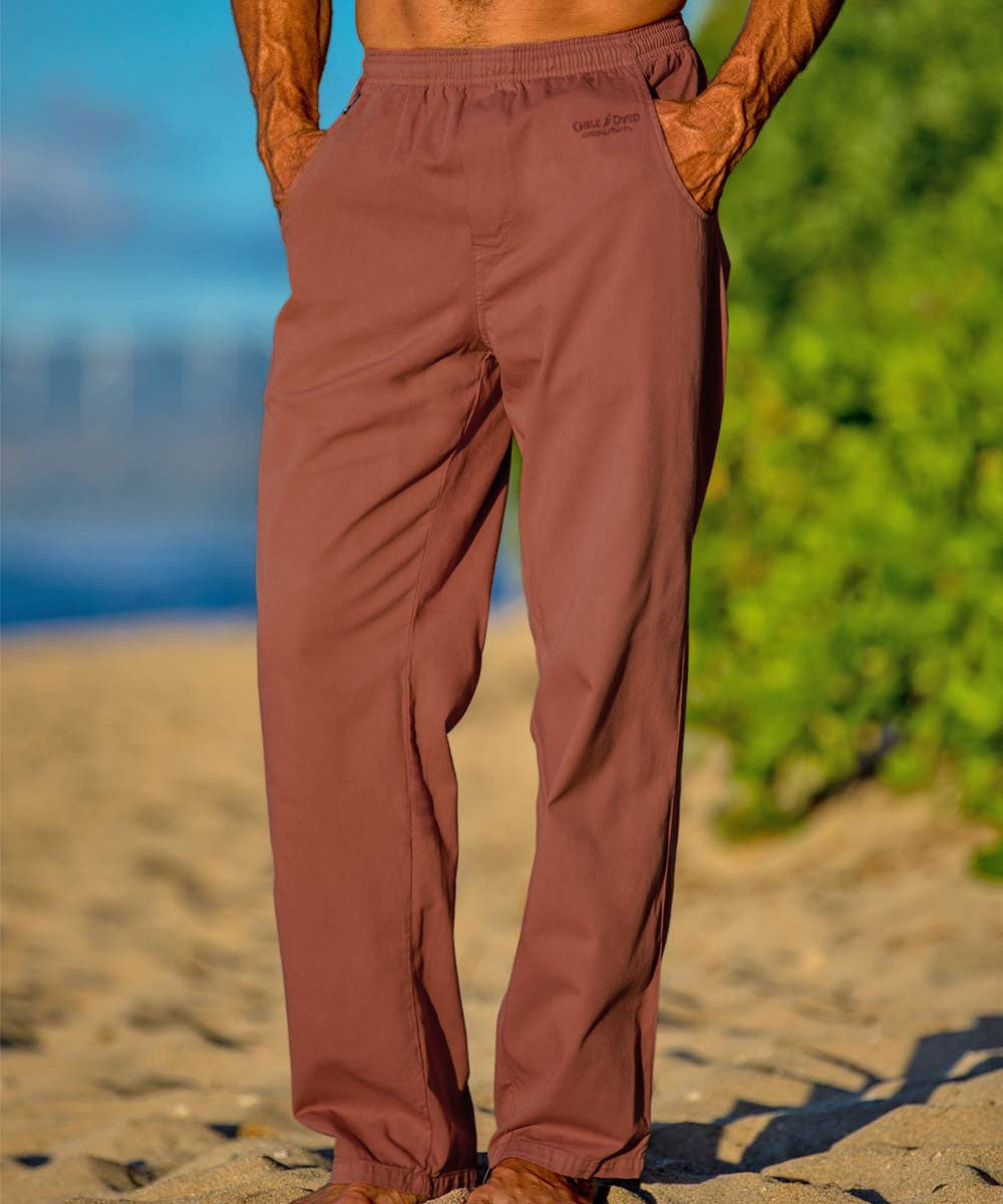 A person standing on a beach wearing rust twill pants with their hands in the pockets. View 2