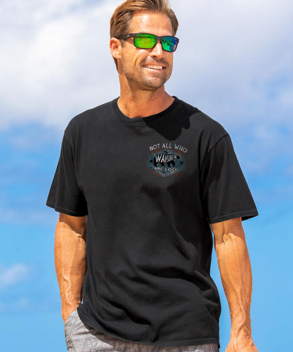 A man wearing a black short sleeve crewneck t-shirt with a graphic text on the front that reads 'Not all who Wander are lost', outdoors. View 3